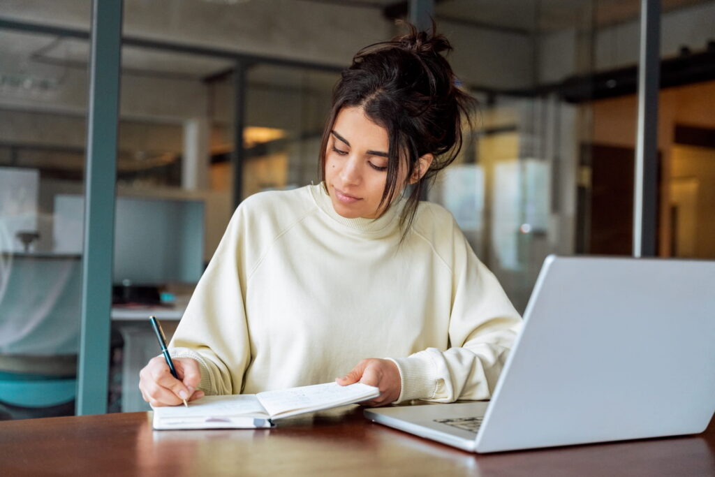 A college-aged woman with dark hair in a bun writes in a notebook at a desk with an open laptop in a modern workspace.